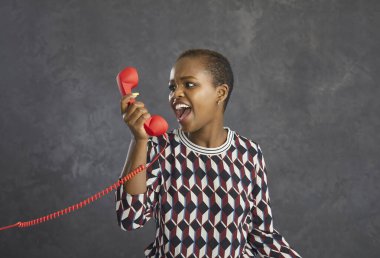 Kızgın Afrikalı Amerikalı kadın telefonda operatörle tartışırken sinirli ve öfkeyle bağırıyor. Gri arka planda duran kırmızı retro telefondan konuşan bir kadın. Olumsuz duygular kavramı.