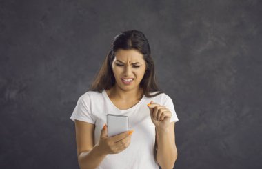 Utanç verici bir şekilde utangaç bir kadın endişeli yüz ifadesiyle cep telefonunun ekranına bakıyor. Genç kadın gri arka planda duruyor ve içerik izlemekten memnun olmadığını ifade ediyor.