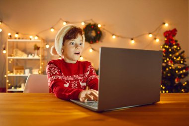 Mutlu sürpriz çocuk evde masada oturuyor ve dizüstü bilgisayar ekranına bakıyor, online aksiyon oyunu oynuyor, internet mağazasında harika bir hediye seçiyor ya da Noel Baba 'dan gelen cevapları okuyor.