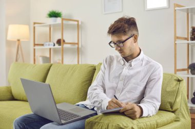 Ciddi bir genç adam çalışmak için dizüstü bilgisayar kullanıyor. Gözlüklü üniversite öğrencisi evdeki kanepede oturuyor, online ders alıyor, bilgisayar ekranına bakıyor ve not defterine not alıyor. Uzaklık öğrenme kavramı