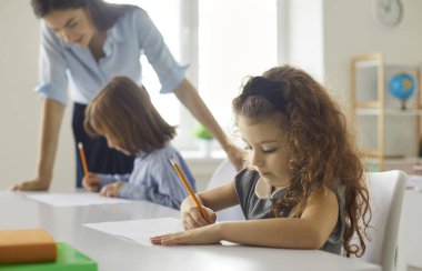 Küçük okul kızının sınıfta bir masada otururken dikkatle yazıp çizdiği yan görüntüler. Kıvırcık kız parlak bir sınıfta sınıf arkadaşlarının ve öğretmenlerinin bulanık geçmişiyle oturuyor.