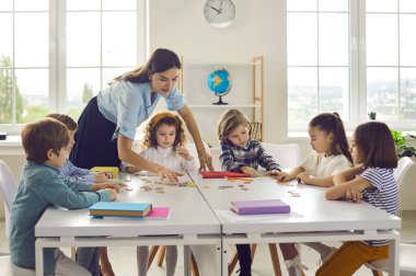 Sınıfta küçük çocuk grubuyla birlikte çalışan genç beyaz kadın öğretmen açıklama yapıyor. Kadın öğretmen ve okul çocukları bulmacaya katılıyor. Anaokulu ya da ilkokul eğitimi ve gelişimi