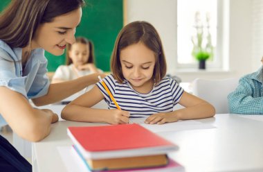 Mutlu öğretmen ilkokulda öğrencilerine yardım ediyor. Küçük bir kız öğrencinin portresini ve görevi doğru şekilde tamamlamaya yardım eden bir bayan öğretmenin portresini kapat. Eğitim ve öğretim kavramı.