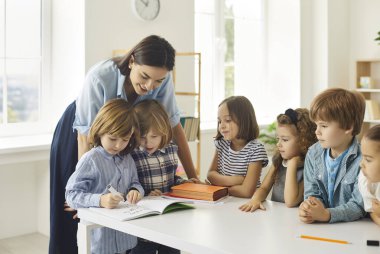 Genç öğretmen not defterlerine yazan mutlu ilkokul öğrencilerine yardım ediyor. Küçük çocuklar sınıfta yeni şeyler öğreniyor. Sınıf arkadaşlarının resim çizmesini izleyen bir grup yetenekli çocuk.