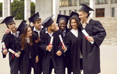 Birlikte üniversite mezuniyetini kutlayan bir grup başarılı çok ırklı öğrenci. Üniversite merdivenlerinin yanında duran akademik önlüklü öğrenciler gülüyor. Eğitim ve mezuniyet kavramı.