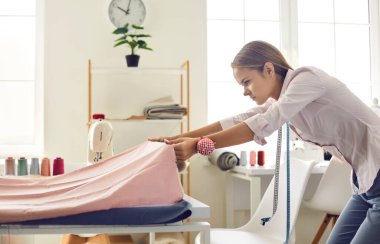 Terzi çalışıyor. Terzi atölyede kıyafet yapıyor. Tekstil terzisi. Ciddi bir kadının yeni tip açık pembe kumaşı masaya yayıp detaylar için hazırlarken yan görüntüsü.