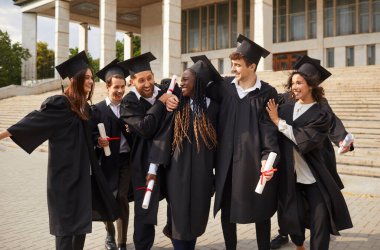 Gülümseyen, mutlu, çok ırklı mezun öğrencilerin üniversite mezuniyet elbisesiyle kucaklaşıp eğlendiği bir grup. Eğitim ve mezuniyet kavramı.