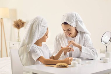 Anne çocuğuna günlük cilt bakımını gösteriyor. Mutlu anne ve güzel kız, yumuşak beyaz başlıklı türbanlar içinde güzellik masasında oturuyor, el kremi sürüyor ve birlikte iyi vakit geçiriyorlar.