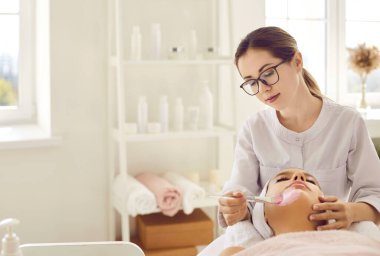 Kadın yüzüne pembe kilden maske takan gözlüklü güzellik uzmanı. Sakin, genç bir bayan modern güzellik salonunda ya da spa merkezinde spa gününde rahatlıyor ve eğleniyor. Boşluk arkaplanı kopyala