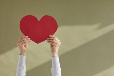 Kalp şeklinde kırmızı bir kağıt tutan kadın elleri, haki yeşili bir arka planda sevgililer günü kartı gibi duruyor. Sevgililer Günü, Dünya Kalp Günü, insan sevgisi, destek, ilgi, minnettarlık, sağlık konseptleri