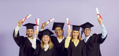 Yüksek öğrenim diplomalarını almaktan mutlu olan neşeli çok ırklı öğrencilerin portresi. Havan topu ve mantoyla mezun olur. Mor arka planda gülümserler. Pankart.