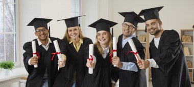 Mezuniyet etkinliğindeki mutlu öğrenciler. Neşeli melez erkek ve kadın koleji ya da üniversite mezunları şapkalar ve bornozlarla sınıfta duruyor, diploma tomarlarını gösteriyor ve kameraya gülümsüyor.