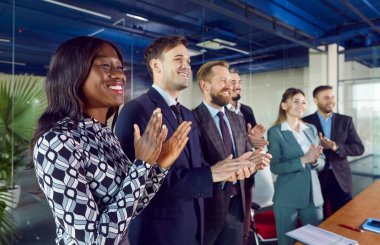 Bir grup farklı genç, sırayla masanın başında ayakta alkışlıyor. Gülümseyen başarılı iş arkadaşları ve şirket çalışanları bir toplantıda veya iş konferansında meslektaşlarını alkışlıyorlar.