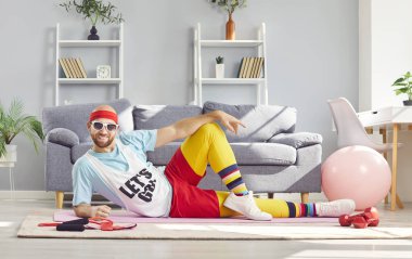 Spor malzemeleriyle çevrili yoga minderinde uzanan renkli spor kıyafetleri içindeki komik müsrif genç adam. Neşeli sporcu antrenman sırasında mola veriyor, oturma odasında poz veriyor ve kameraya bakıyor..