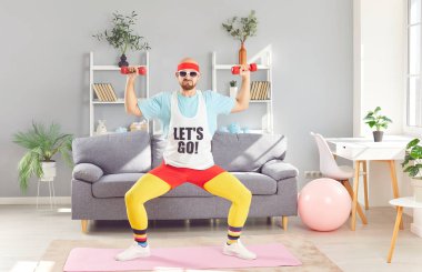 Komik kıyafetli, güneş gözlüklü ciddi kel bir adam ev spor salonunda dambıl egzersizi ve çömelme hareketleri yapıyor. Fitness eğitimi ve antrenman rutini mizah ve spora bağlılığın bir karışımını gösteriyor..
