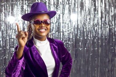 Parlak gümüş arka planda kadife takım elbiseli mutlu ve şık esmer tenli genç kadın. Mor elbiseli, şapkalı ve gözlüklü komik kadın parti fikri için parmağını kaldırıyor. Pankart.