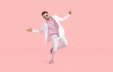 Pastel pembe arka planda izole edilmiş beyaz takım elbiseli mutlu genç adamın portresi. Modern moda takım elbiseli ve gözlüklü kaygısız bir adamın stüdyoda eğlendiği uzun bir çekim.