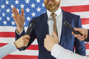 Takım elbiseli Amerikalı erkek politikacı, gazete muhabirleri ve televizyon gazetecileri tarafından sorularla bombardımana tutulurken birkaç mikrofona konuşuyor. Basın toplantısı, ABD rali kampanyası konsepti