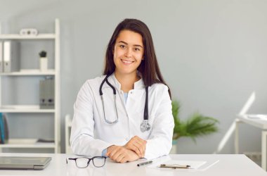 Kadın doktor görüntülü arama yapıyor ve hastaya online danışma modunda yardım sağlıyor. Ofisinde web kamerasının önünde oturan beyaz ceketli, arkadaş canlısı genç bir kadının portresi..