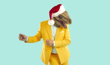 Komik bir Noel kostümü giymiş mutlu bir adamın portresi. Parlak sarı takım elbiseli, dinazor maskeli, kırmızı Xmas şapkalı, turkuaz bir arka planda dans eden garip bir adam.