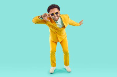 Ziyaretçi işaretini gösteren komik bir adam stüdyodaki açık mavi arka planda sana bakıyor. Parlak sarı takım elbiseli ve güneş gözlüklü şık bir adam gülümsüyor. Tam uzunluk.