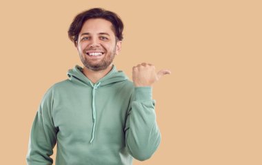 Dost canlısı, mutlu, esmer, yeşil tişörtlü, başparmağıyla bej arka plandaki fotokopi alanını gösteren ve kameraya bakan genç adam. Reklam, pazarlama, satış konsepti.
