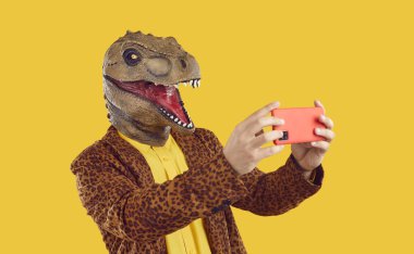 Lastik dinozor maskeli tuhaf komik adam sarı arka planda akıllı telefondan selfie çekiyor. Leopar desenli ceket ve sarı tişört giyiyor. Reklam afişi, funky adamla pazarlama.