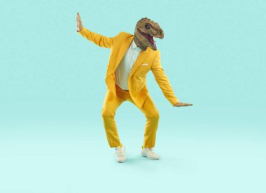 Dinozor kostümü giymiş komik, neşeli bir adam dans ediyor ve eğleniyor. Parlak sarı takım elbiseli mutlu bir adam ve turkuaz mavisi stüdyo arka planında dans eden komik bir dinozor maskesi. Parti konsepti