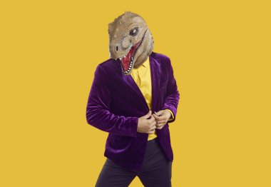 Dinozor kostümlü bir adam moda stüdyosunda poz veriyor. Mor kadife takım elbiseli komik erkek modelin portresi ve sarı arka planda izole bir şekilde duran Cadılar Bayramı dinozor maskesi. Çılgın moda konsepti