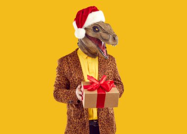 Mutlu, neşeli, heyecanlı, aptal Tyrannosaurus dinozor maskesi takan, kırmızı Noel Baba şapkası takan, elinde Noel hediyesi olan, sarı arka planda izole edilmiş bir leopar ceketli genç adam.