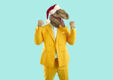 Noel kostüm yarışmasında garip kılık değiştirmiş garip bir adam zaferi kutluyor. Sarı takım elbiseli, Tyrannosaurus dinozor maskeli, Xmas şapkalı, turkuaz arka planda duran mutlu bir adam.