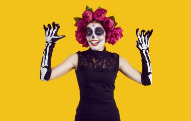 Kafatası şekeri makyajlı mutlu bir kadının portresi, İskelet eldivenler, kafasında çiçekler, elleri korkutan ve sarı arka planda sırıtan. Cadılar Bayramı ve Ölülerin Günü konsepti.