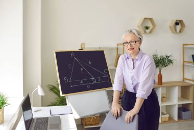 Modern eğitim. Üniversite öğrencilerine uzaktan eğitim veren yüksek matematik öğretmeni bir bayanın portresi. Dost canlısı bir kadın sınıfta dizüstü bilgisayar ve tahtanın yanında. Çevrimiçi eğitim kavramı.