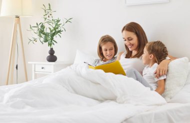 Genç mutlu anne yatak odasında beyaz battaniyenin altında oturup kitap okuyor ya da çocuklarına evde uyudukları için peri kuyruğu anlatıyor. Aile eğlencesi ve uyku zamanı konsepti.
