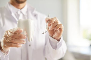 Diş hekimliği kliniğinde elinde diş aynası ve dişle duran beyaz üniformalı genç bir dişçinin portresi. Diş sağlığı ve hijyen konsepti.