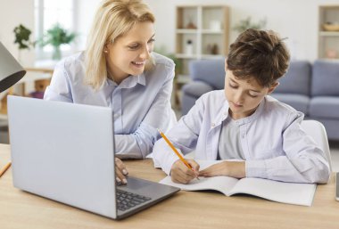 Anne oğluna evde ödevlerinde yardımcı oluyor, online eğitim için dizüstü bilgisayar kullanıyor. Sahne, ebeveyn ve çocuk arasındaki destek ilişkisini yansıtıyor, öğrenme ve desteği vurguluyor..