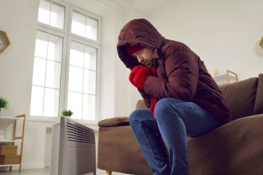 Mutsuz genç adam evinde üşümüş bir şekilde kanepede oturuyor. Kışlık ceket giyen titreyen adam, örgü şapka ve eldiven giyip kendini elektrikli ısıtıcıda ısıtıyor. Soğuk kış mevsiminde evde üşüyor.