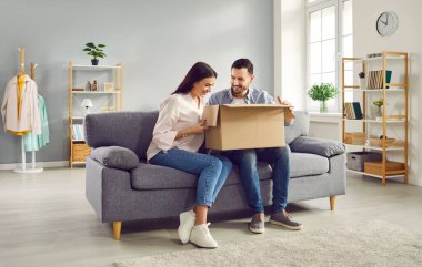 Happy young joyful couple unpacking parcel at home, sitting in the living room on sofa and smiling looking into open cardboard box. Delivery, online shopping, receive the parcel concept.