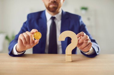 İş adamı elinde bir bitcoin ve bir soru işareti tutuyor, bu da dijital finans, para ve ticarette risk ve belirsizliği simgeliyor. Kripto para, iş ve mali yatırım kavramı.