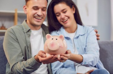 Mutlu genç bir çiftin portresi. Aile bütçelerini planlıyorlar. Kanepede oturuyorlar ve ellerinde domuz kumbarası tutuyorlar. Yatırımlar ve finansal özgürlük kavramı.