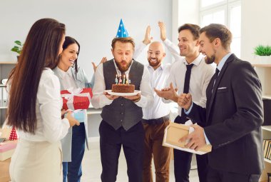 Bir grup gülümseyen iş adamı iş arkadaşlarını doğum günüyle kutluyorlar. Doğum günü pastası tutan erkek çalışan işyerindeki mumları üflüyor. Şirket partisi