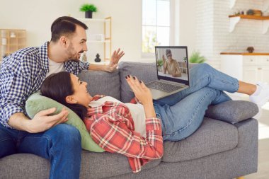 Genç, gülümseyen bir çift evde kanepede dinleniyor ve bilgisayardan yetişkin bir anneyle sohbet ediyorlar. Erkek ve kadın el sallayarak yaşlı ebeveynleri selamlıyor..