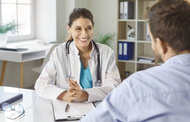 Erkek hasta, ofisteki masada oturan gülümseyen genç bir doktoru ziyaret ediyor. Doktor klinikte muayene sırasında bir adama danışıyor. Tıp konsepti.