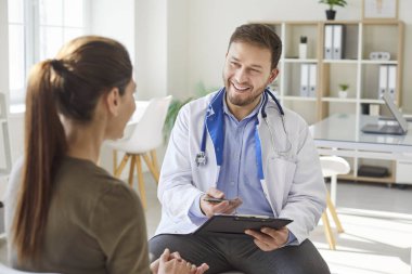 Dost canlısı genç erkek doktor terapistle konuşan bir kadın klinikteki tıbbi muayene sırasında iş yerindeki randevusuyla birlikte rapor tutuyor. Tıp konsepti.