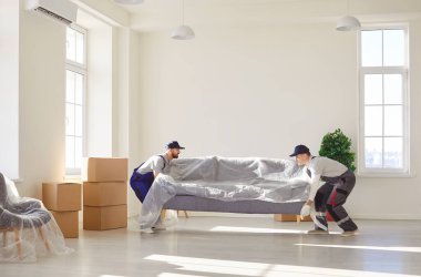 Parlak geniş bir odada plastiğe sarılı ağır gri kanepe taşıyan üniformalı iki profesyonel yükleyici. Mobilyaların taşınması, taşınma işlemleri yeni modern apartmanda işçiler tarafından yürütülüyor..