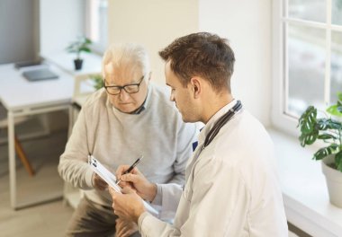 Tıp bürosundaki yaşlı, gri saçlı bir hastanın portresi elinde randevu ile rapor tutan ve klinikteki tıbbi muayene sırasında danışmanlık yapan erkek doktoru dinliyor..