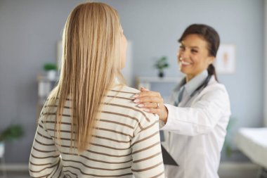 Neşeli doktor, hastasına sağlığındaki olumlu değişiklikler hakkında bilgi veriyor. Nazikçe omzuna dokunuyor. Doktor, kameraya arkası dönük duran kadın hastaya pozitiflik ve destek gösteriyor..