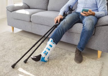 Yaralı bacağı olan, ortopedik çizme giyen, koltukta oturan, yanında koltuk değnekleri olan, akıllı telefon kullanan, ayak travmasından sonra dinlenen bir adam..