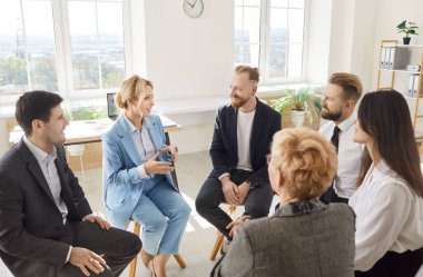 Bir grup iş adamı bir çemberin içinde oturmuş modern ofiste iş projesini tartışıyorlar. İş antrenörü, takım lideri veya koç şirket çalışanlarıyla kalkınma stratejisi hakkında konuşuyor.