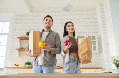 Mutlu genç çift, ellerinde alışveriş poşetleri ve taze sebzelerle mutfakta dikiliyorlar. Gülümseyen adam ve kadın paketleri açıyor, sağlıklı yemek hazırlıyor, evde birlikte vakit geçiriyorlar. Düşük açı